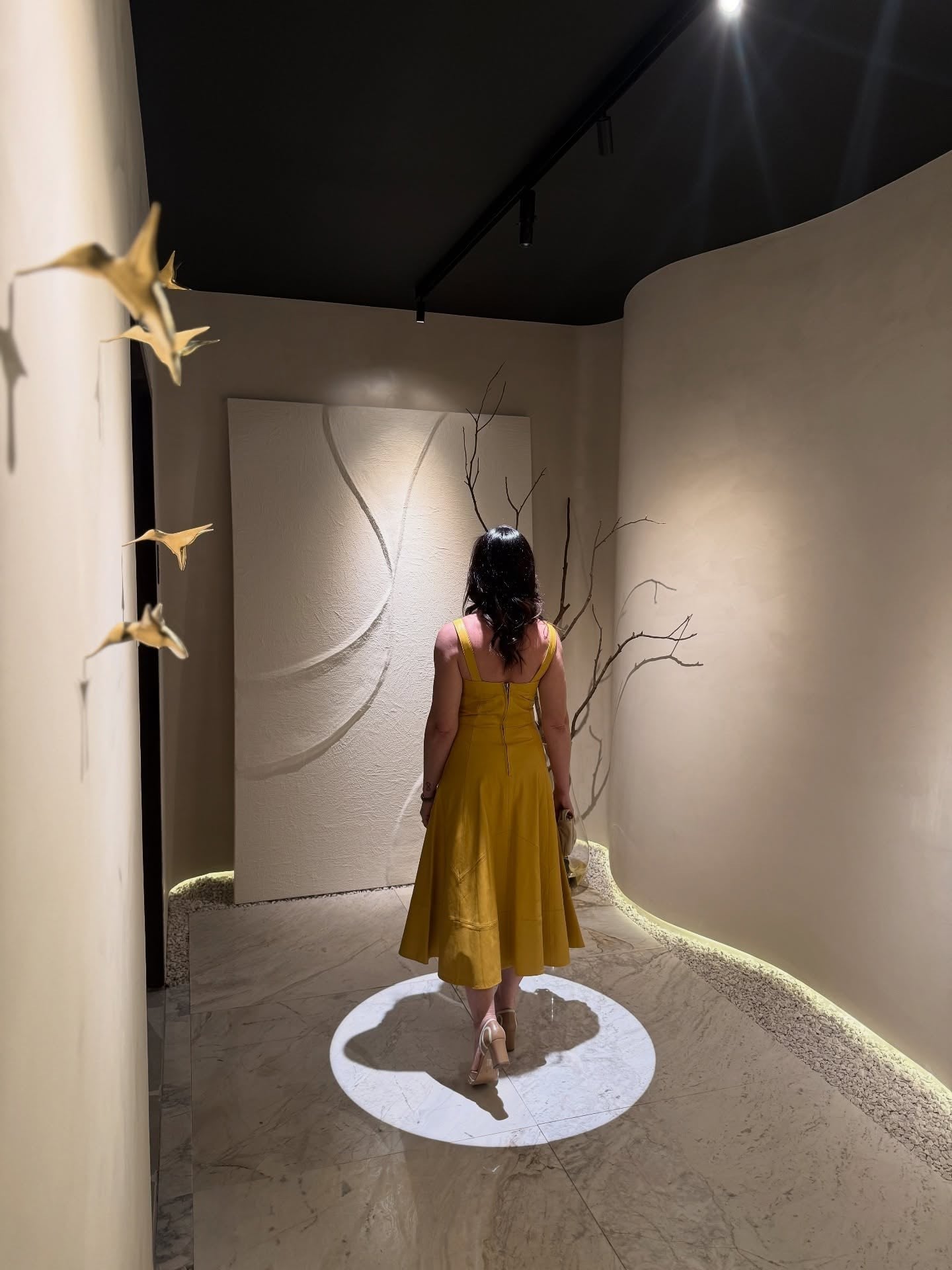 Carolina Arruda, de vestido amarelo, de costas, caminhando pela galeria sobre o círculo de luz