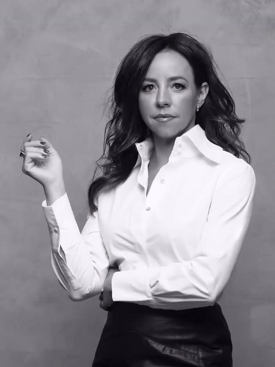 Retrato em preto e branco de Carolina Arruda, de camisa branca, diante de parede clara