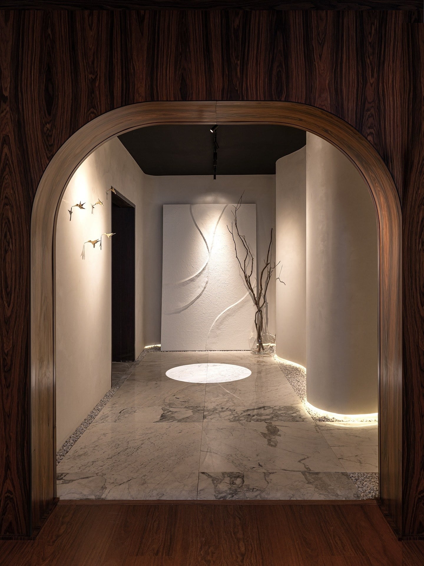 Arco de madeira escura emoldurando a galeria clara, com escultura na parede e luz circular no piso