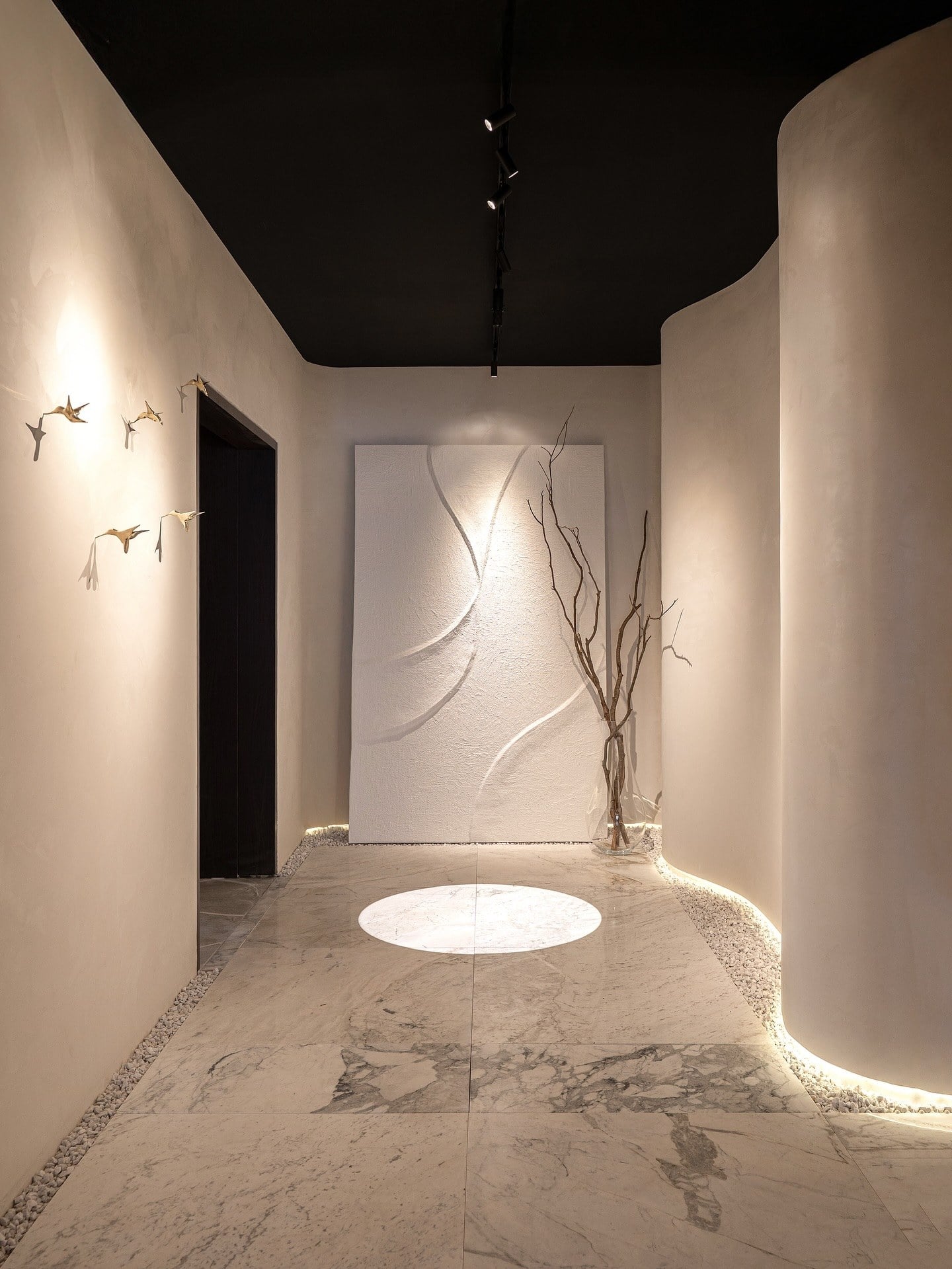 Galeria de paredes curvas com teto escuro, painel esculpido e galhos secos sob a luz